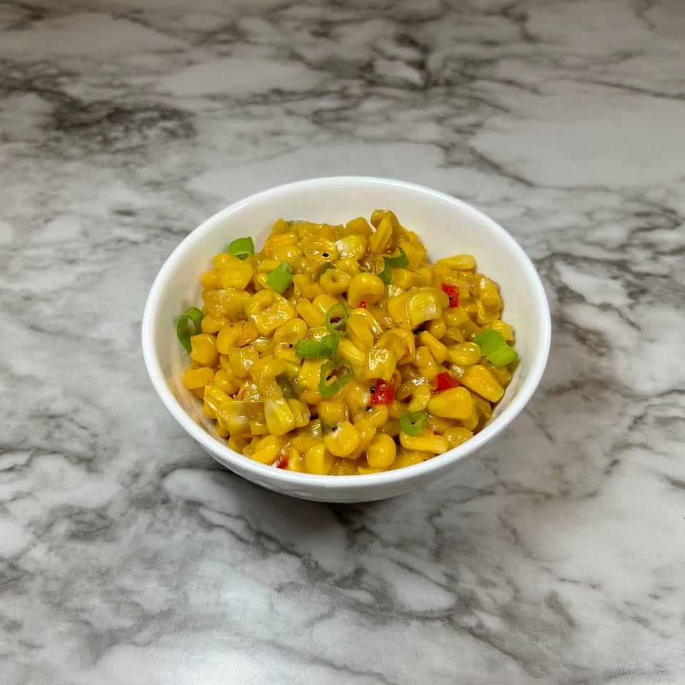 A white bowl of creamy yellow corn, garnished with green onions and red peppers, on a marble surface.