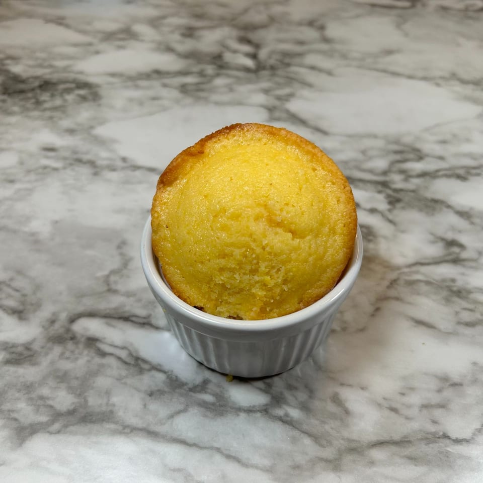 A golden-brown cornbread muffin in a white ramekin, centered on a marble surface.